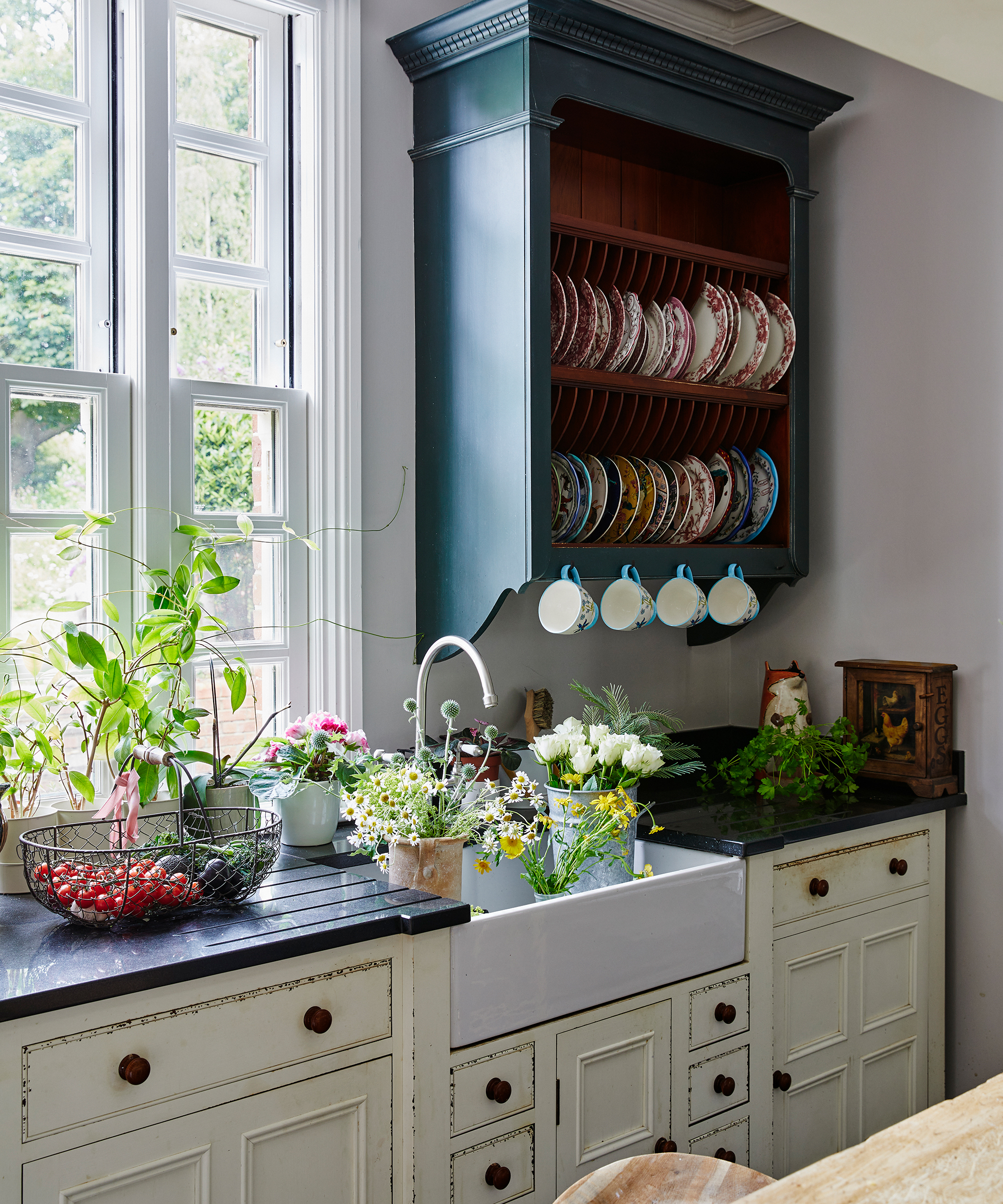 sink in country kitchen by window filled with flowers with wall plate rack beside