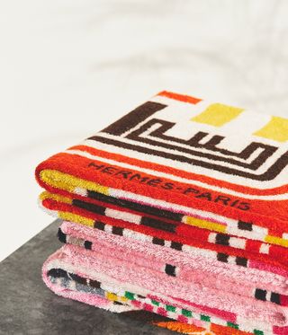 View of a pile of folded, multicoloured and patterned Hermès towels on a grey surface pictured against a light coloured background
