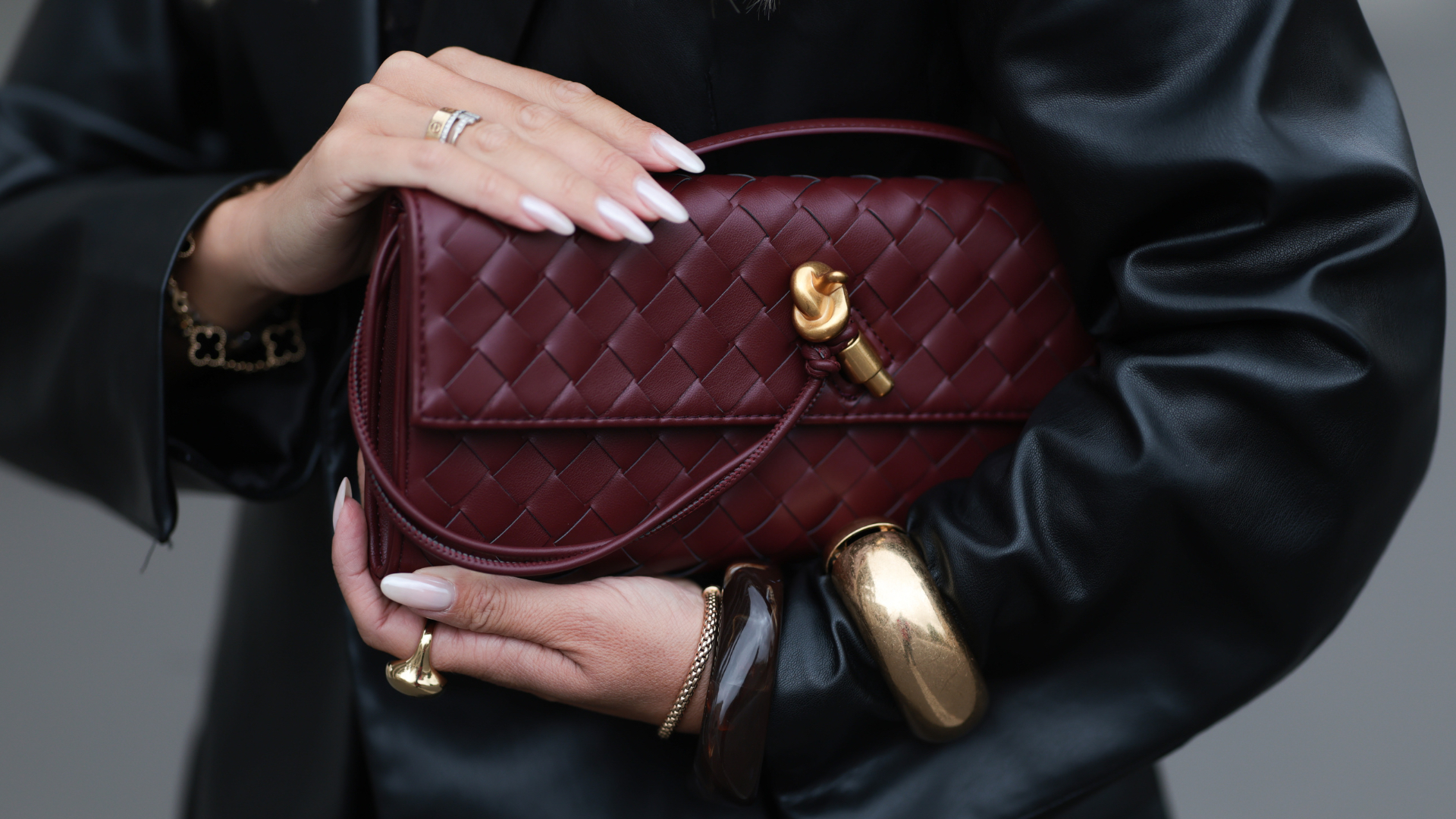 Hard Biab Olga Liber with long, softly waved blonde hair worn in a center part, seen wearing a black satin lace camisole paired with loose charcoal silk trousers and an oversized black leather blazer. She completes the look with a gold-buckled YSL belt, oversized Prada shades, and a burgundy woven leather clutch