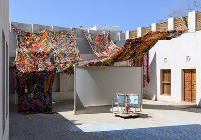 An open courtyard space with a brown door and brown window on the right featuring light blue merry go-round with 2 cseats, colourful knitted ropes formed into large blanket like hanging items