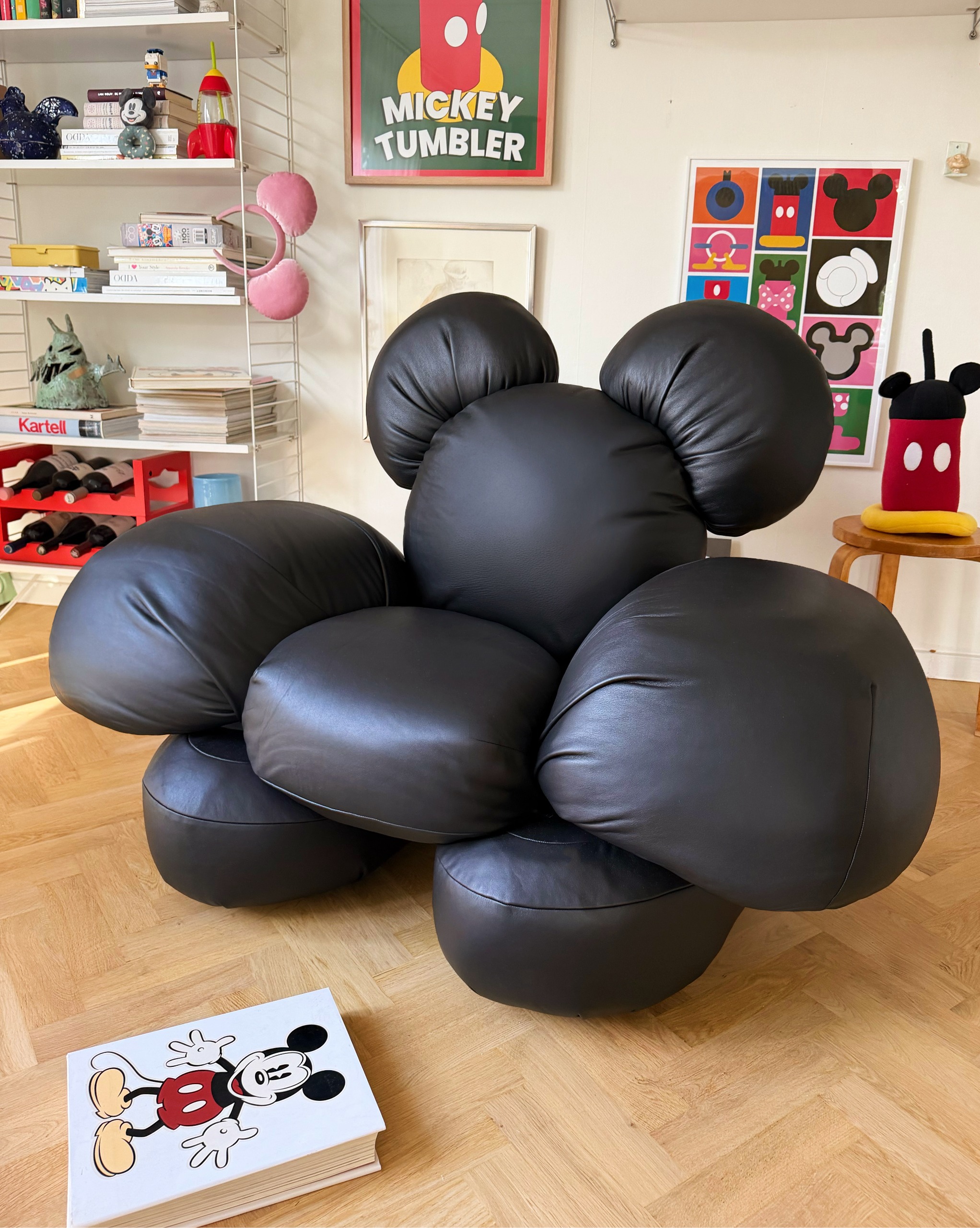 A mouse-shaped, eight-piece black leather sofa, styled in a wooden floored room with colorful prints and a bookcase in white-painted metal bookcase, styled in front of a Mickey book.