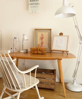 A retro looking wooden desk with easels on it, a white rocking chair with striped grey and white cushion, and floor lamp next to it