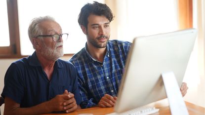 A young man teaches an older man something on a computer.