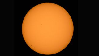 a large orange sphere the sun, is at the center of the image. There us a tiny black dot to the lower left of the sun, this is Mercury. A large sunspot is also visible towards the upper center of the sun.