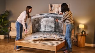 Two women unpack the Helix Twilight Mattress in a box, still compressed in plastic, onto a bed frame.