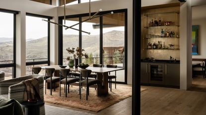 A modern dining space with a home bar and sweeping mountain views through large blame-framed floor to ceiling windows