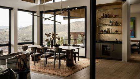 A modern dining space with a home bar and sweeping mountain views through large blame-framed floor to ceiling windows