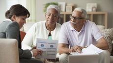 A husband and wife who are retired having a financial consultation with an adviser at their home, looking at financial sheets and a laptop