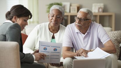 A husband and wife who are retired having a financial consultation with an adviser at their home, looking at financial sheets and a laptop