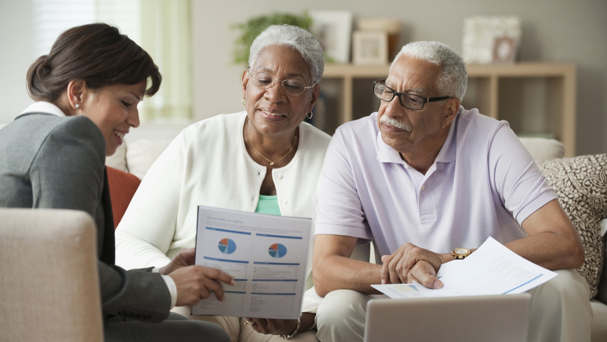 A husband and wife who are retired having a financial consultation with an adviser at their home, looking at financial sheets and a laptop