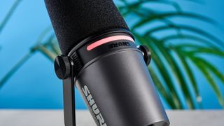 the shure mv7i, a black microphone with a black muff, touch LED pad, XLR connection and TRS input and USB-C input at the rear, photograhped against a blue tom's guide background with a pair of over-ear wired headphones