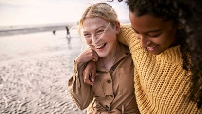 two friends with arms around each other, laughing on a beach