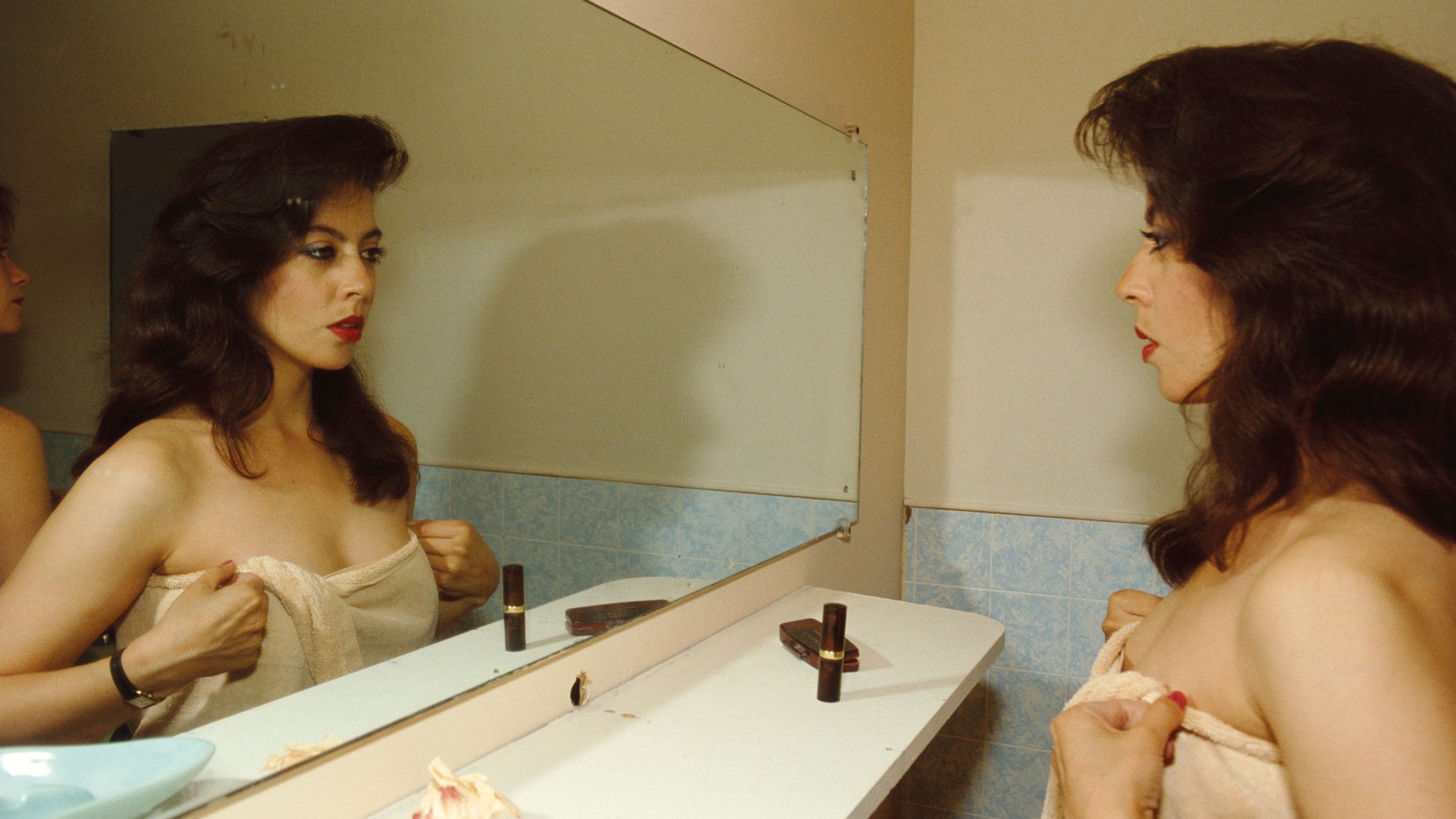 A woman with voluminous dark hair and red lipstick gazes into a bathroom mirror while holding a towel around her body.