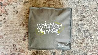 YnM Grey weighted blanket carry case on a floral rug. Carry case says 'weighted blankets' and 'feels like a hug'