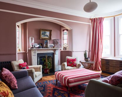 Ottoman ideas. Pink painted living room space with bright bay window, navy sofa, two cream striped armchairs, positioned around a pink striped ottoman, curtains with matching pink striped pattern, wooden floor with multi colored pattern rug, traditional marble fireplace with mantel decorated with vases, photographs and decorative glass objects