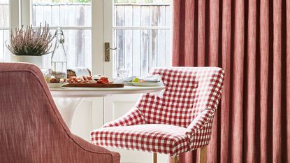 A round white dining table with two pink chairs, one checked, one plain. Large white doors behind them with a long pink curtain. 