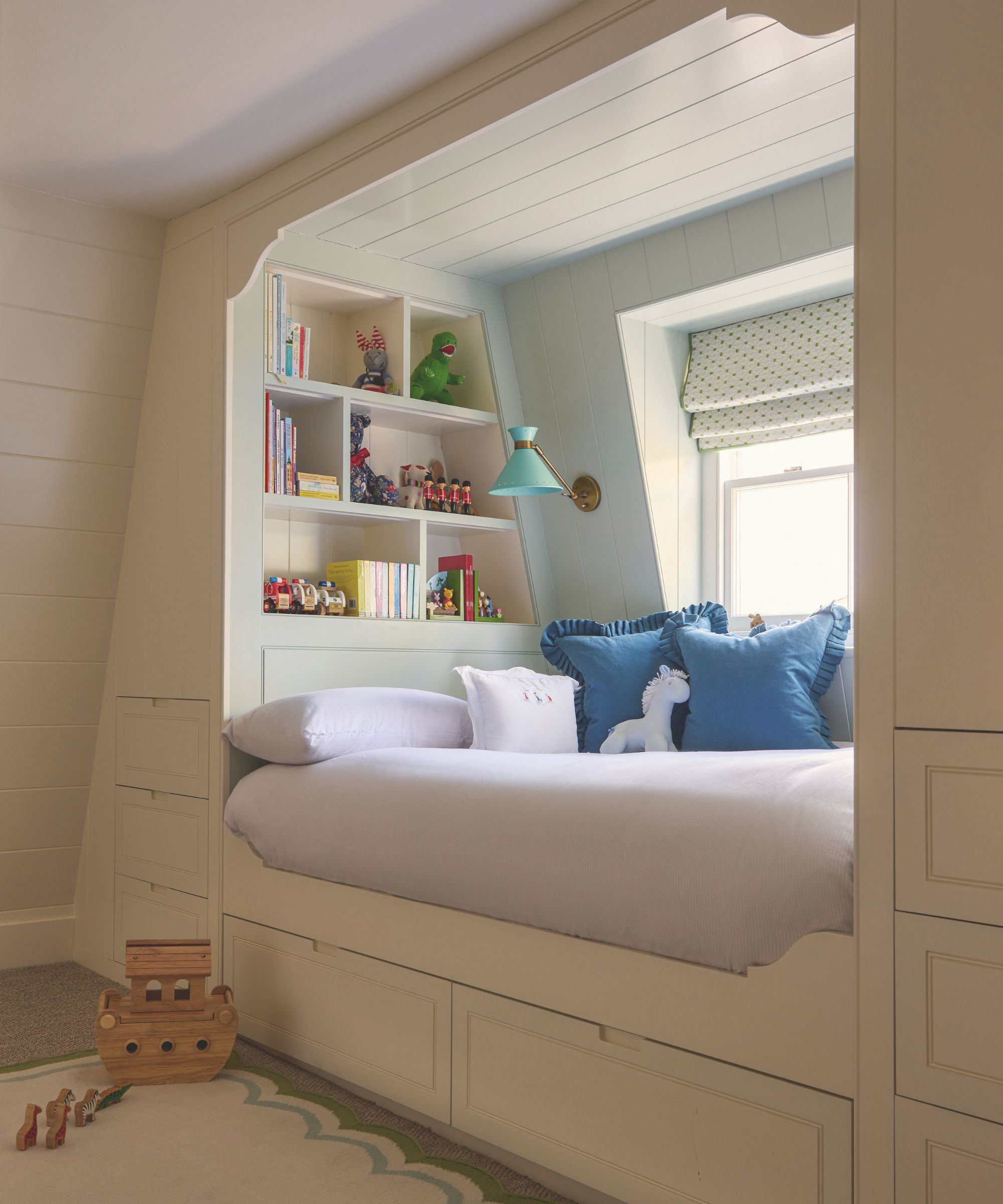 A custom-built children&amp;rsquo;s alcove bed tucked into a cream paneled wall. The cozy nook includes built-in bookshelves filled with toys, blue ruffled cushions, a teal reading lamp, and integrated storage drawers underneath.
