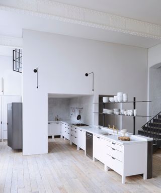 An all-white kitchen features a surprising wall in the middle of the design. Metal planks feature as kitchen cabinets, balancing plates.