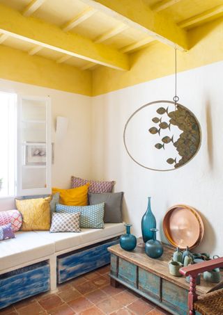 A living room with a yellow painted ceiling, tiles on the floor, and an ottoman bench by the window with multi-colored cushions, and a wooden cabinet with vases and mugs on top