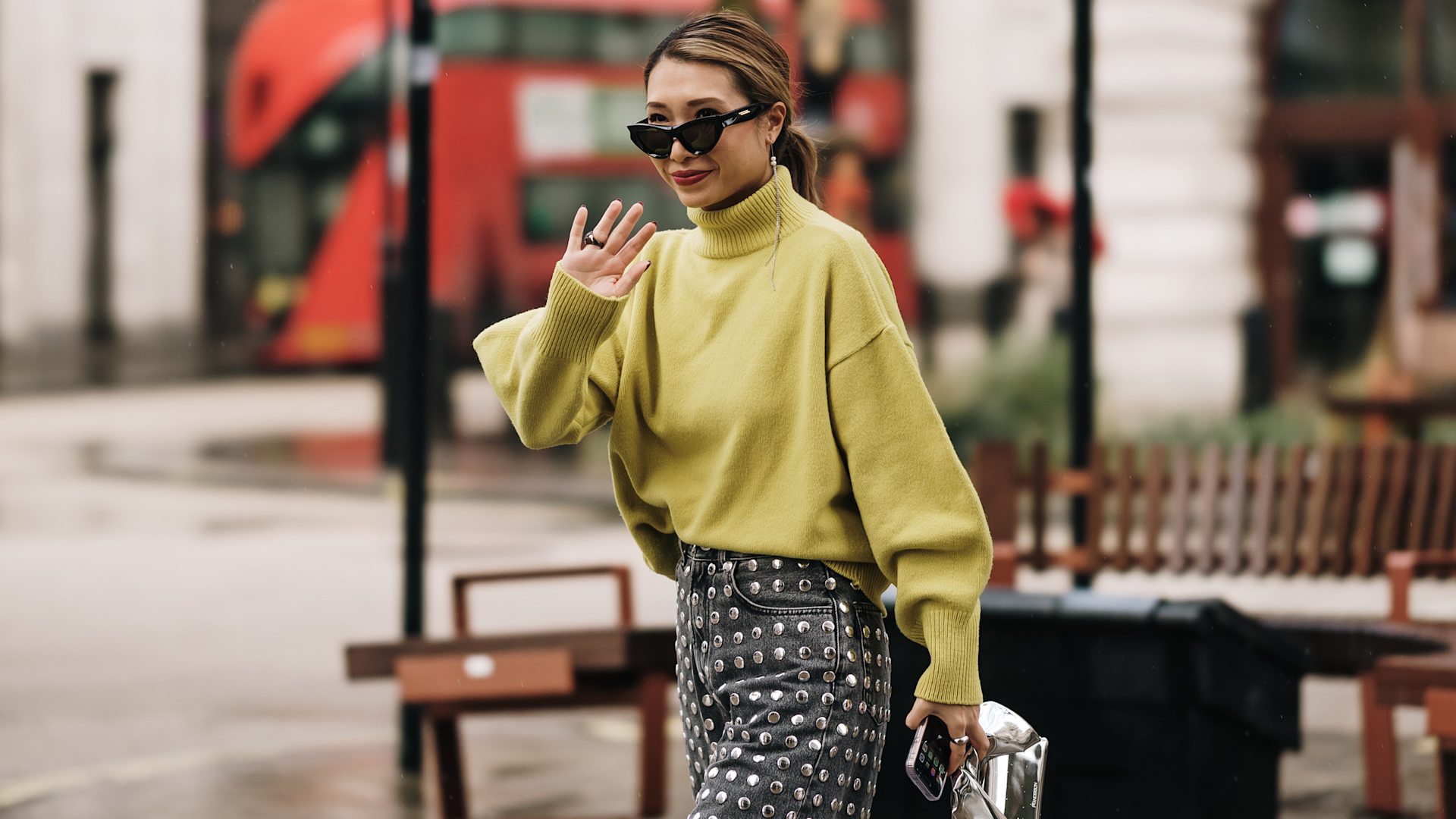 fashion week attendee wears sunglasses, green sweater, and studded jeans, waving 