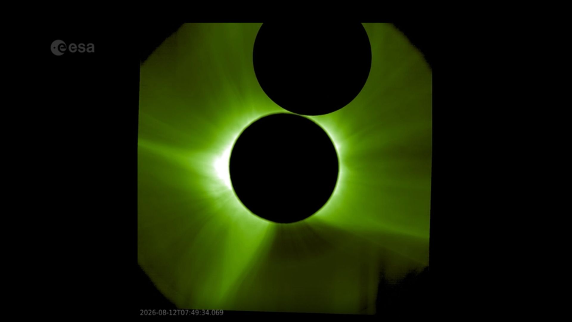 An image of the sun captured by a spacecraft. The sun's corona shines green from behind the black circle of a sunshield, which hides the sun's surface. The moon's silhouette can be seen above.