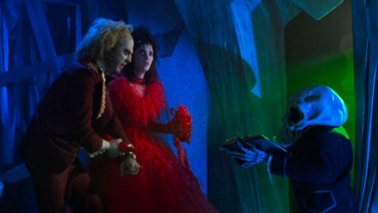 Michael Keaton como Beetlejuice e Winona Ryder em um grande vestido vermelho enquanto Lydia fala com o pequeno alienígena em Beetlejuice.