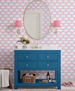 A pink small bathroom with tulip patterned wallpaper, a round gold mirror, two pink wall sconces, and a dark blue vanity unit