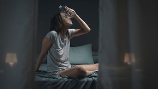 A woman sits up in bed sweating in the middle of the night, trying to cool herself down by holding a bottle of water to her forehead.