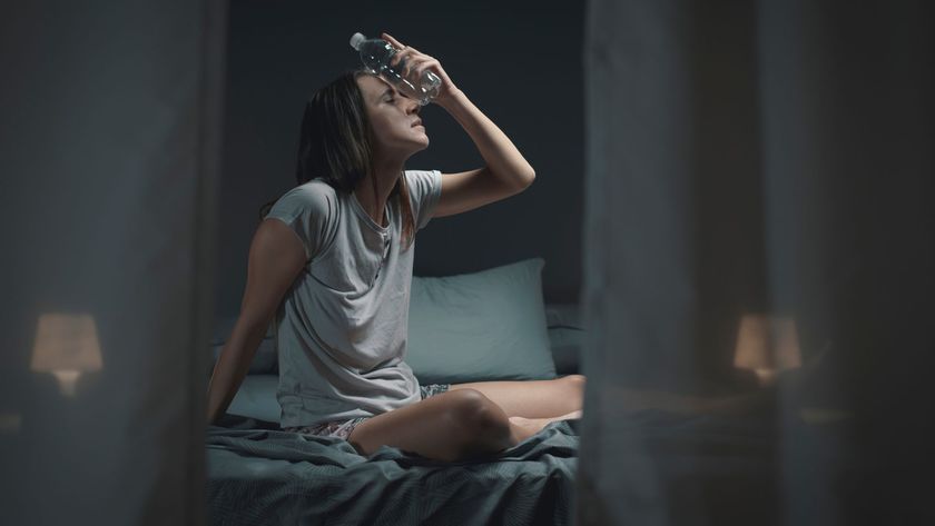 A woman sits up in bed sweating in the middle of the night, trying to cool herself down by holding a bottle of water to her forehead.