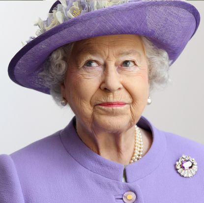 Queen Elizabeth wearing a purple coat and jeweled brooch with a purple floral-topped hat