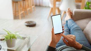 A woman lays on a sofa relaxing and using a phone while a robot vacuum cleans the floor