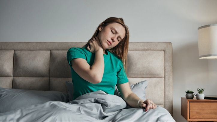 A woman wearing green tshirt sitting in bed with her bed tilted and her hand on her neck.