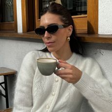 Woman wearing white sezane cardigan and black sunglasses, holding up coffee cup.