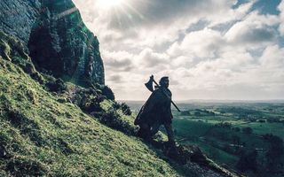 Dev Patel as Gawain walks down a rocky hillside carrying an ax in The Green Knight