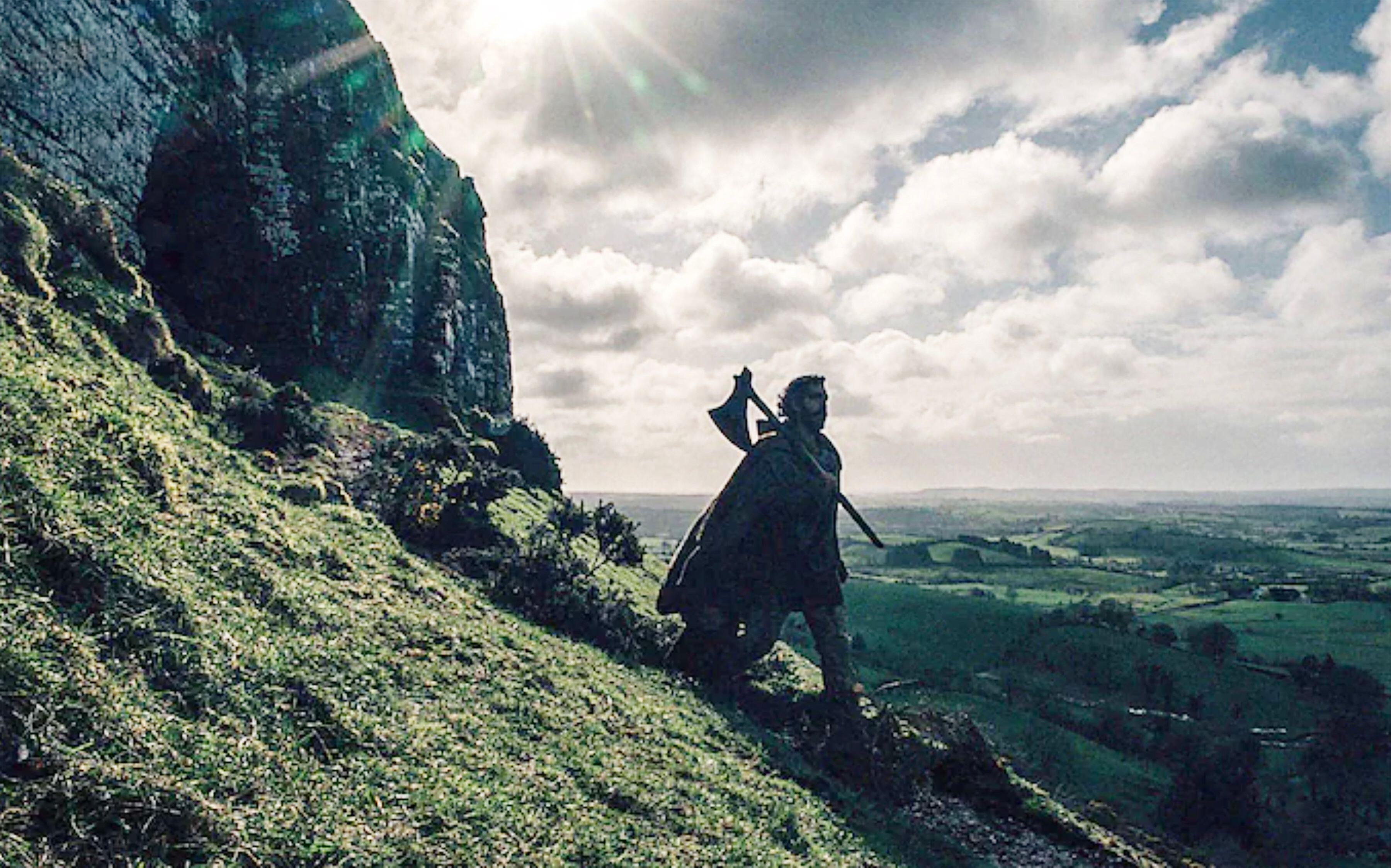 Dev Patel mientras Gawain camina por una ladera rocosa llevando un hacha en The Green Knight