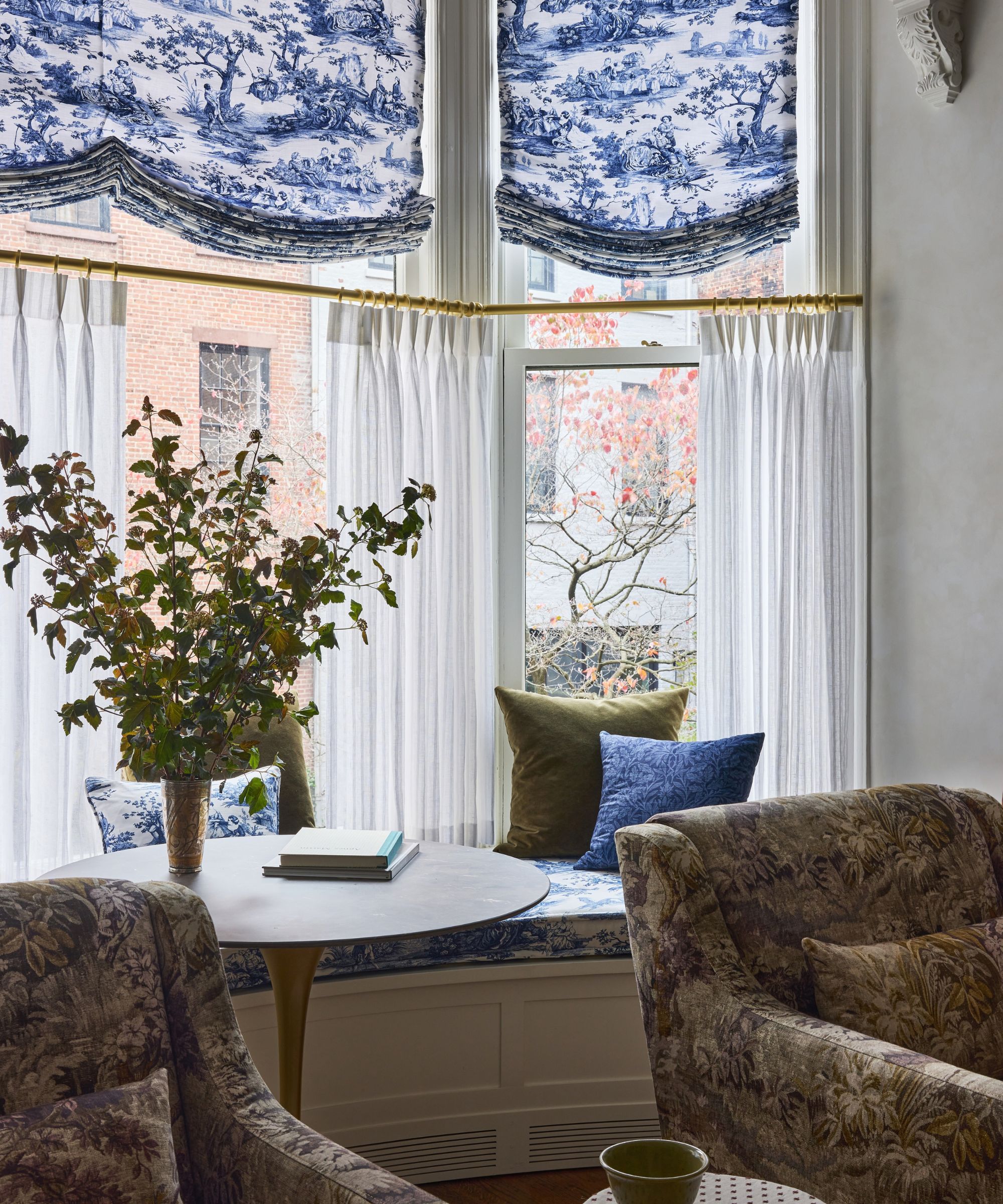 A window seat with large windows and sheer cafe curtains, toile de jouy blinds, a built-in bench with matching cushions, a round wooden table with a vase of flowers, and two brown armchairs.