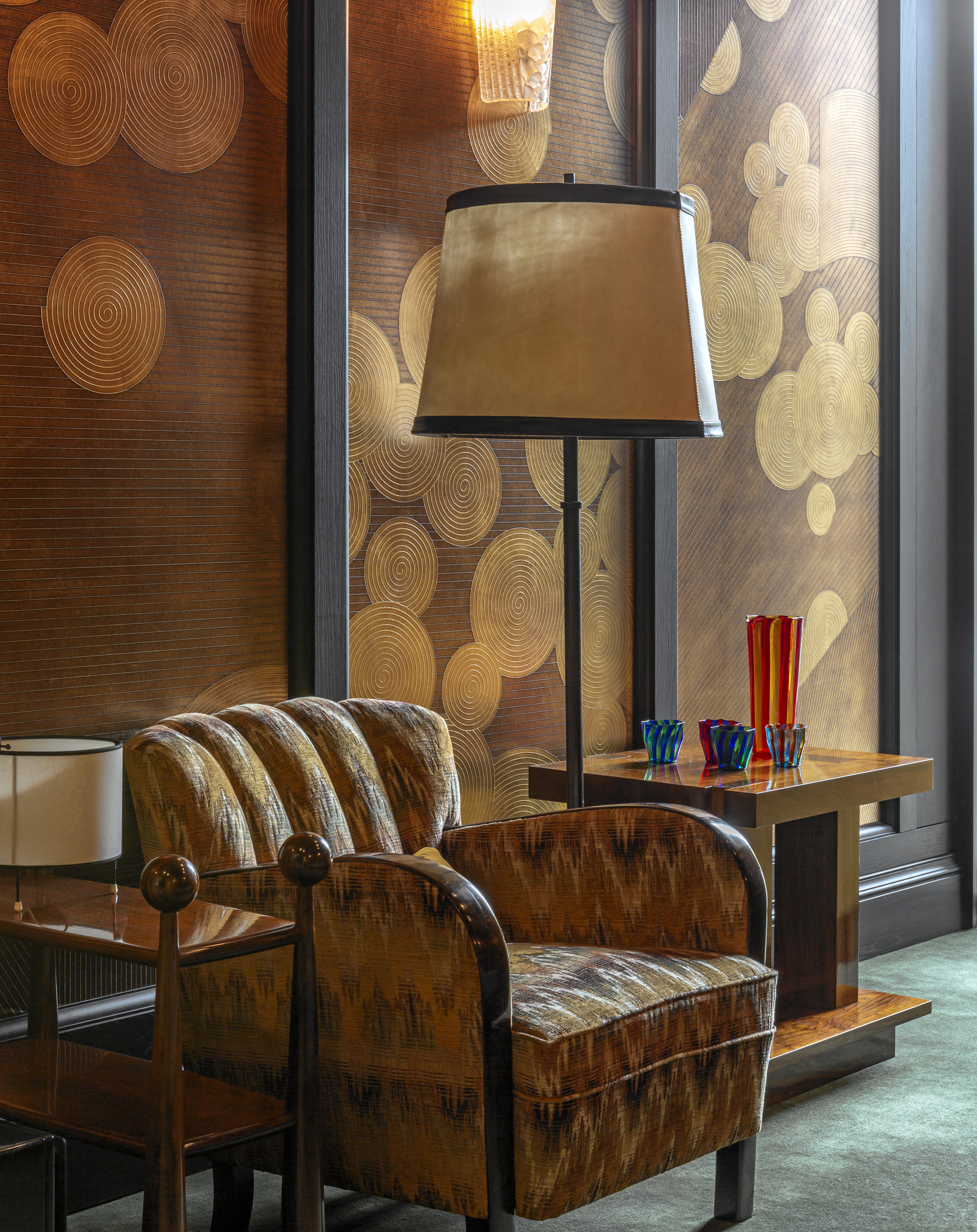 art deco style space with gold patterned wallpaper, art deco style upholstered chair, floor lamp, side table with glass vases styled on it, and green carpet