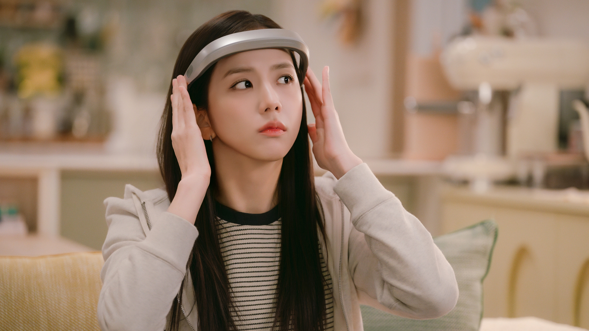 A woman (Jisoo as Seo Mi-rae) holds a headband-style device to her forehead as she sits on a couch in a living room, in the Netflix K-drama 'Boyfriend on Demand.'