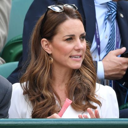 Kate Middleton wearing a white dress and sunglasses on top of her head holding a pink tube of lip gloss while sitting in front of a green metal railing at Wimbledon 