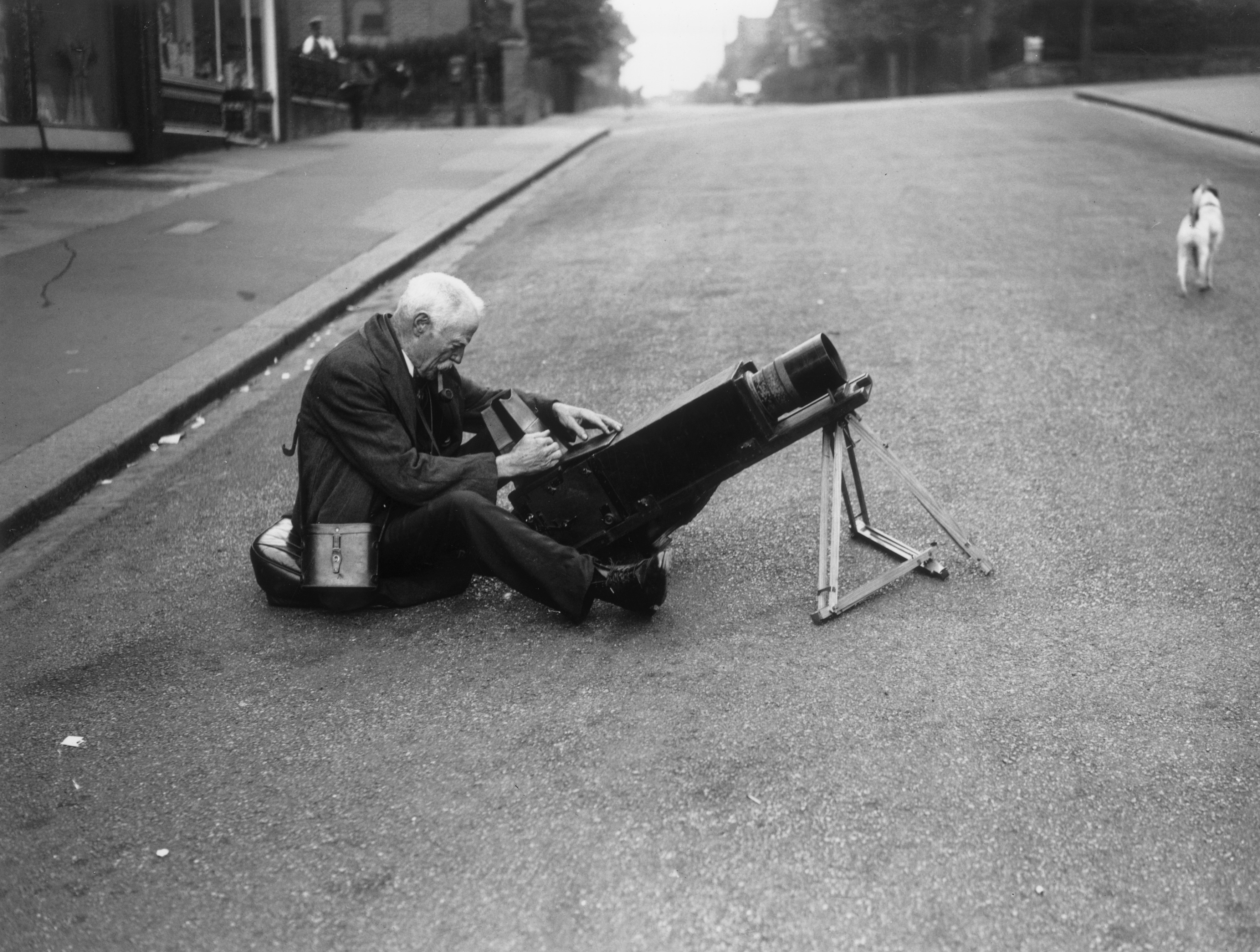 A man sits in the middle of the road with his photography kit and a pipe in his mouth, a small dog is running up the road into the distance.