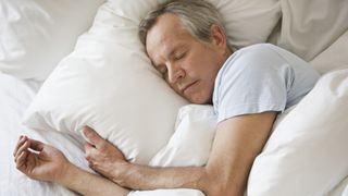 How to sleep better: Middle aged man with gray hair asleep in bed covered by a white comforter