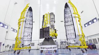 A wrapped up satellite sits on a metal cart next to two halves of a large cylindrical case, both also on carts in a cleanroom