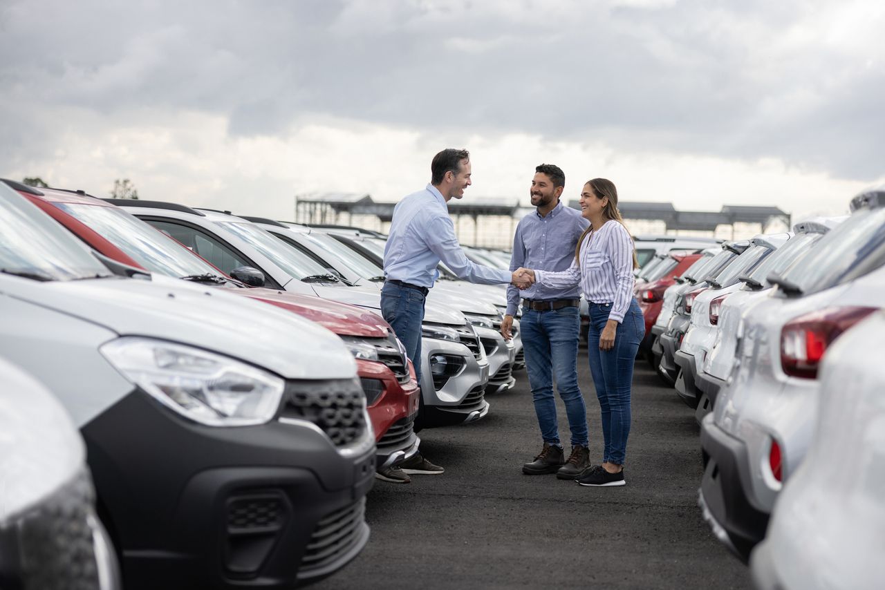 A couple shakes hands with a car dealer in a parking lot full of cars to buy or lease.