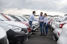 A couple shakes hands with a car dealer in a parking lot full of cars to buy or lease.