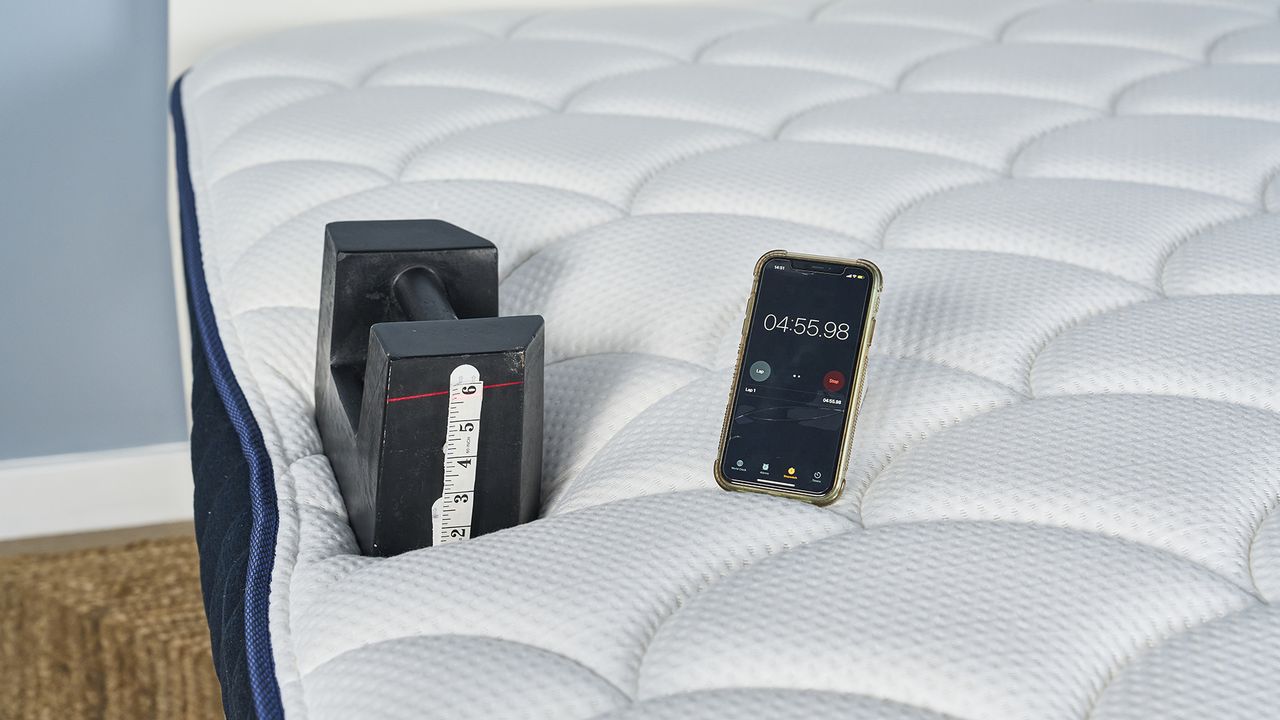 A black 25kg weight 3" away from the side edge of the DreamCloud Classic. A red laser line shows it has sank over 5" and a phone next to the weight shows almost 5 minutes has passed.
