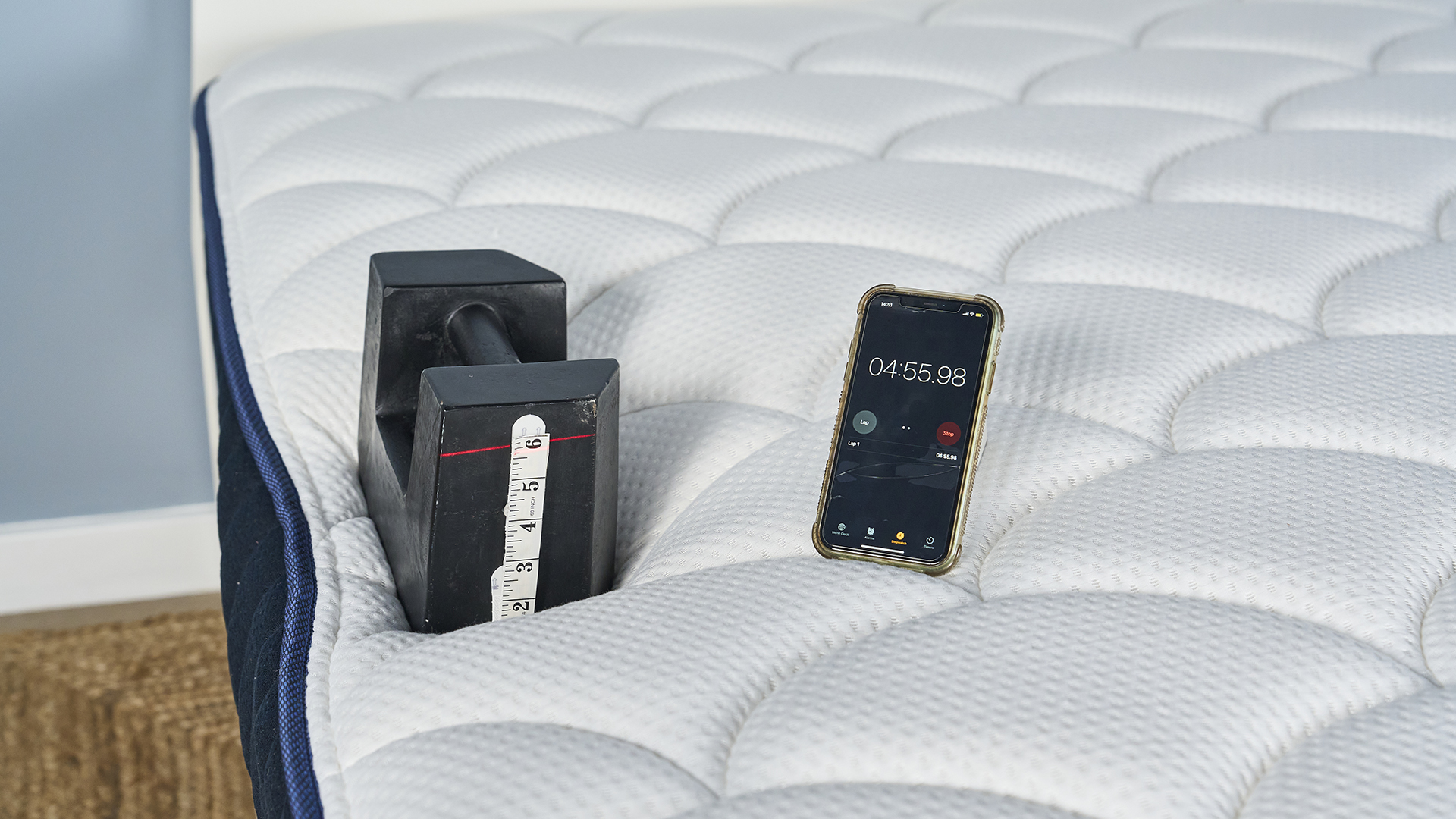 A black 25kg weight 3" away from the side edge of the DreamCloud Classic. A red laser line shows it has sank over 5" and a phone next to the weight shows almost 5 minutes has passed.