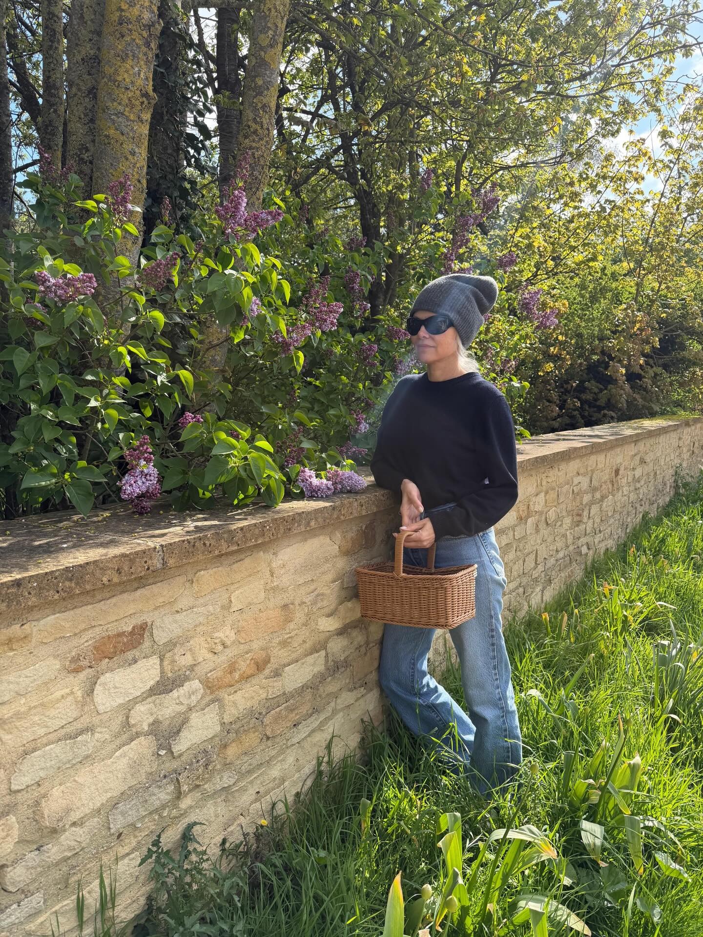 Image of Pamela Anderson in a garden wearing jeans with a navy jumper, beaning and black sunglasses.