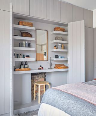 A bedroom with large cupboard doors open to show a dressing table within, woodwork painted pale grey. Built in wall of cupboards and storage. Master bedroom