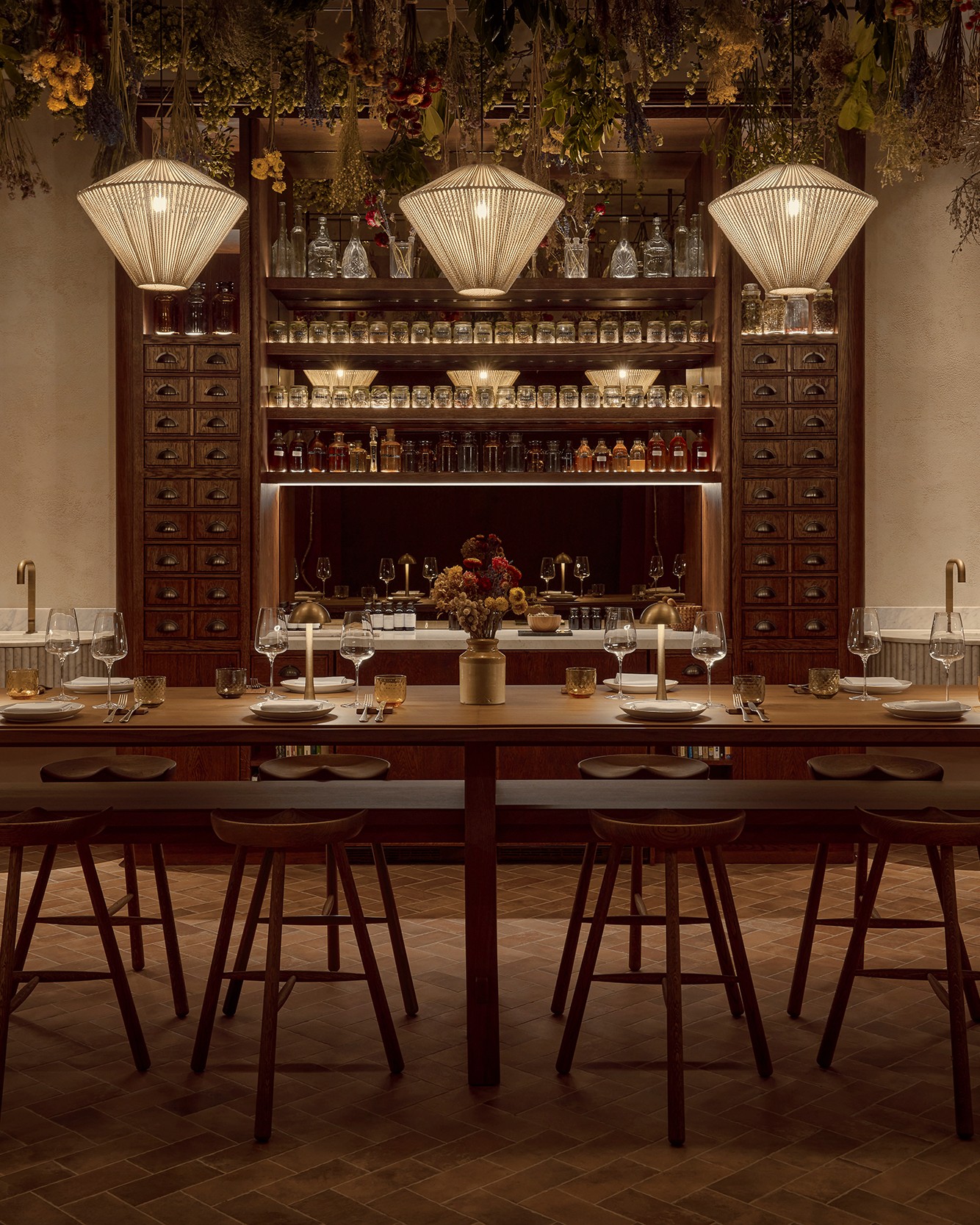 A woody dining room and bar with an apothecary-style cabinet filled with spices and herbs kept in jars, dried herbs pending from the ceilings, and pendant beige lamps.