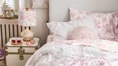 A dorm room with pink and white floral sheets, a white headboard, and a pink table lamp on a cream nightstand